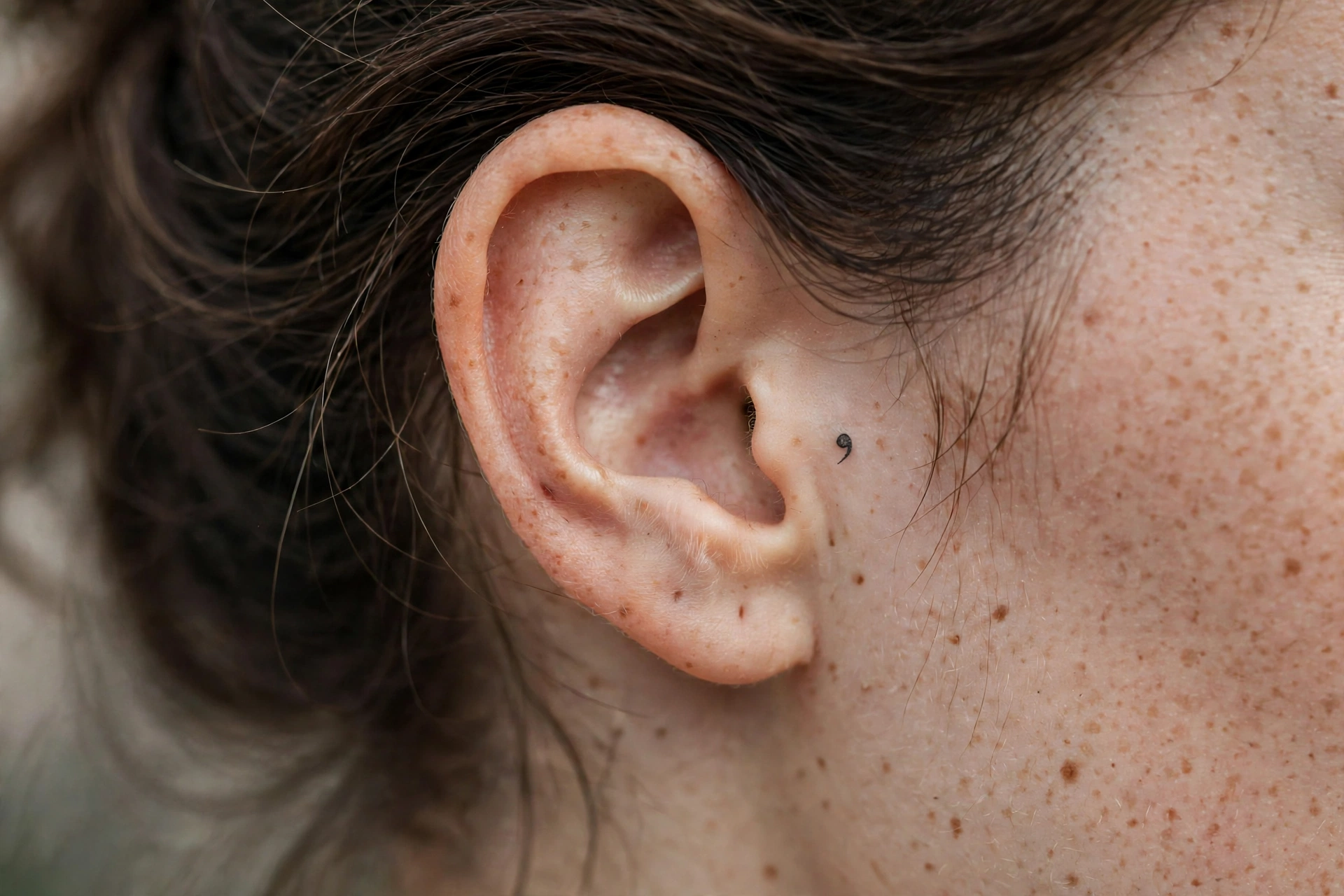 Tiny minimalist semicolon tattoo behind an ear, close-up